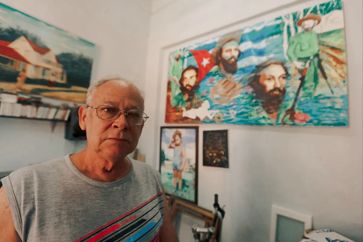 Senior man in an artistic workshop surrounded by colorful paintings, wearing eyeglasses and a gray shirt.