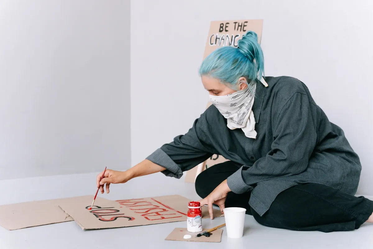 Woman with blue hair paints protest signs emphasizing social activism.