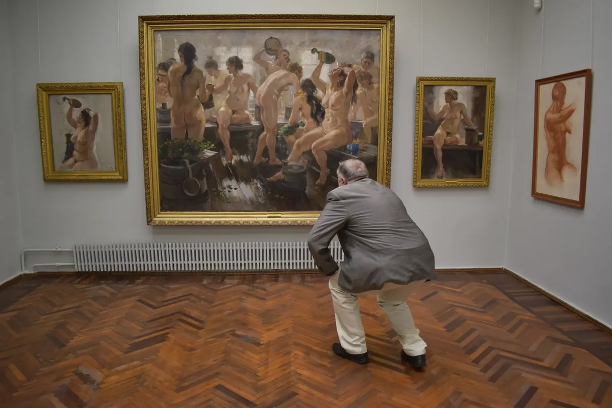 A man closely examines paintings in an art gallery, featuring nude art.