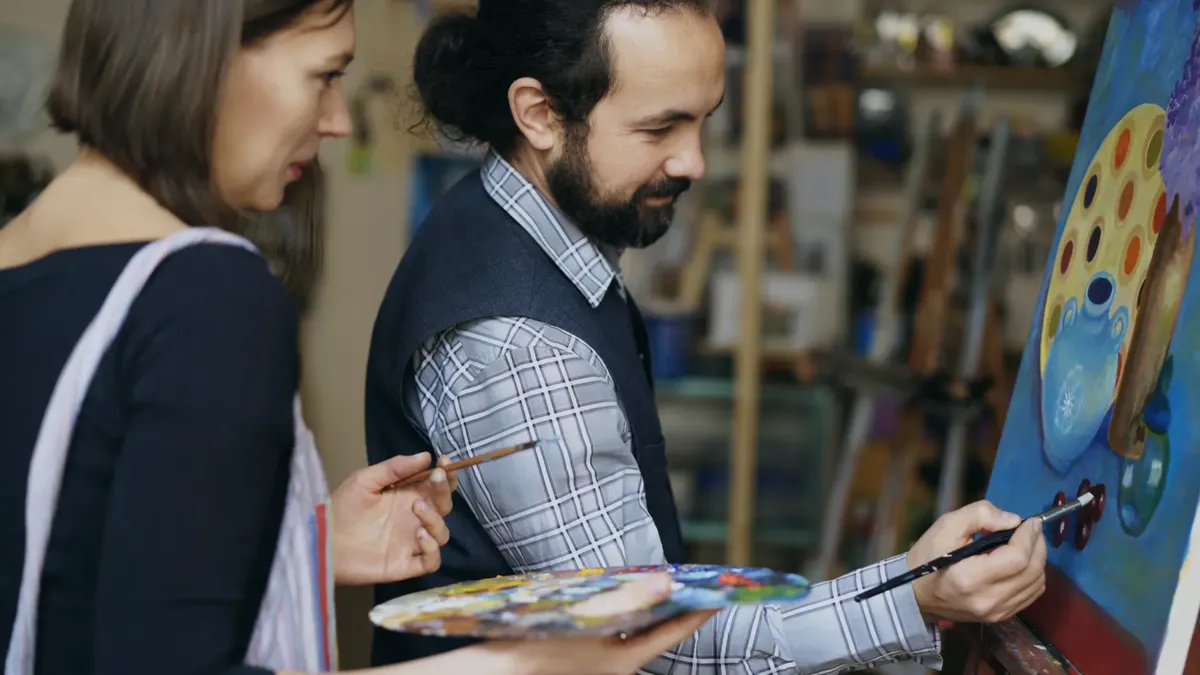 Two adult artists painting together on a canvas in an art studio environment.