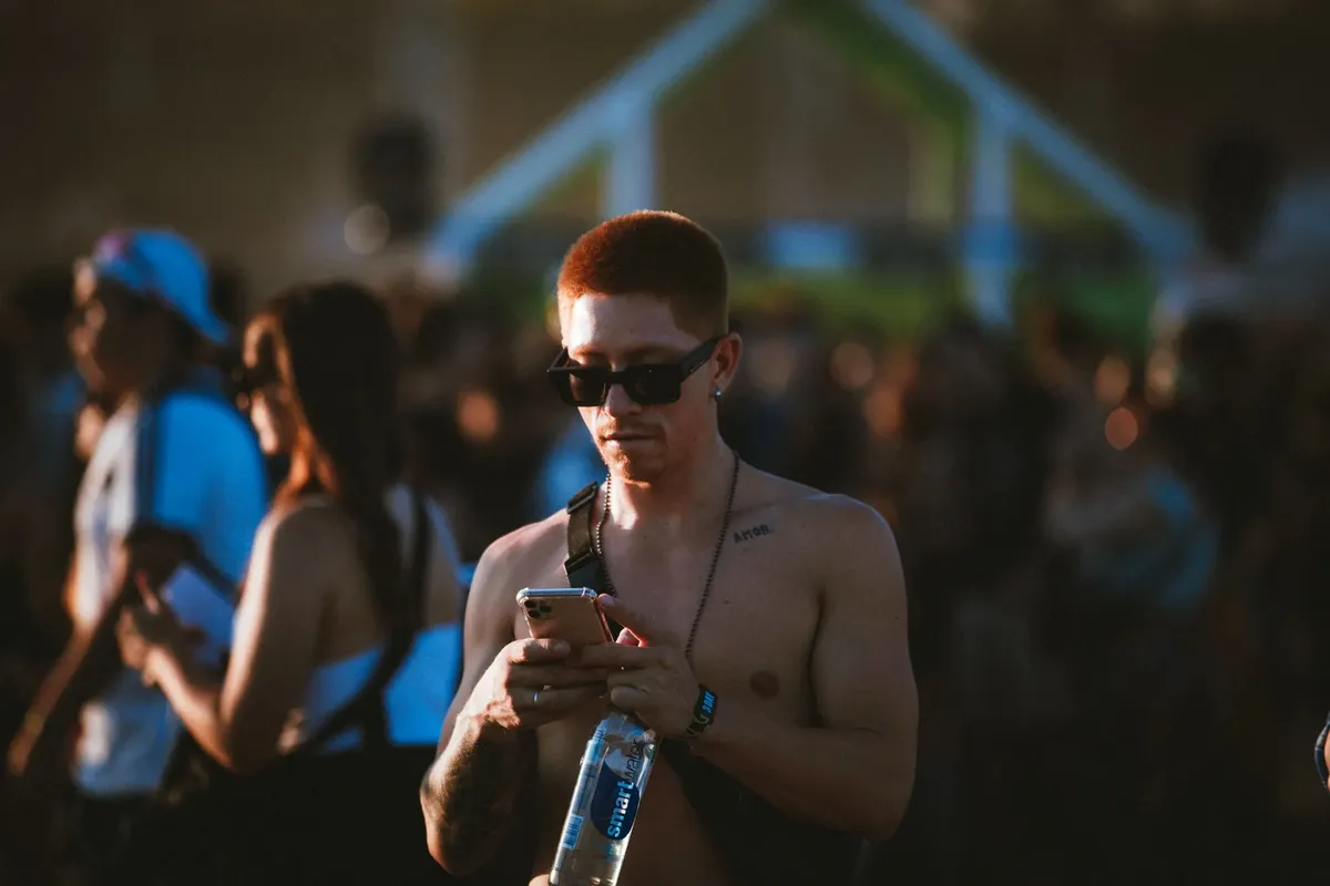 Shirtless man wearing sunglasses at a lively outdoor music festival, checking phone.