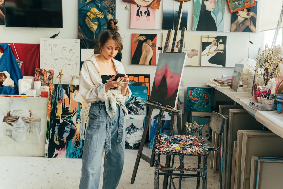 A female artist in a dynamic studio, surrounded by colorful paintings and tools, uses a smartphone.